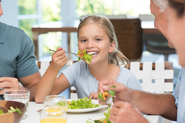 Cheerful little girl enjoying lunch with her family. Portrait of a happy smiling daughter eating salad for lunch and looking at camera. Happy female child having lunch with extended family at home.